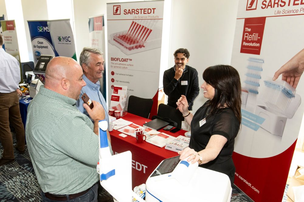 Two male attendees talking to two females at a conference exhibition stand standing beside a table covered in front of a scientific computer sensor and some company promotional information leaflets.
